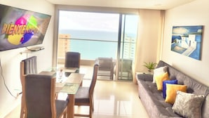Una sala de estar moderna con comedor, un sofá con cojines, un televisor de pantalla plana y un balcón con vista al mar.