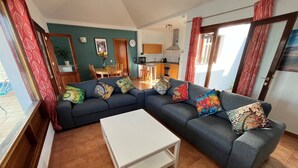 Een woonkamer met een grijze hoekbank, een witte salontafel en kleurrijke kussens. Er is een eethoek met houten meubels en een keuken op de achtergrond.