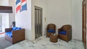 Een hal met een bank, twee rieten stoelen, een bijzettafeltje, een vlag en een lift.
