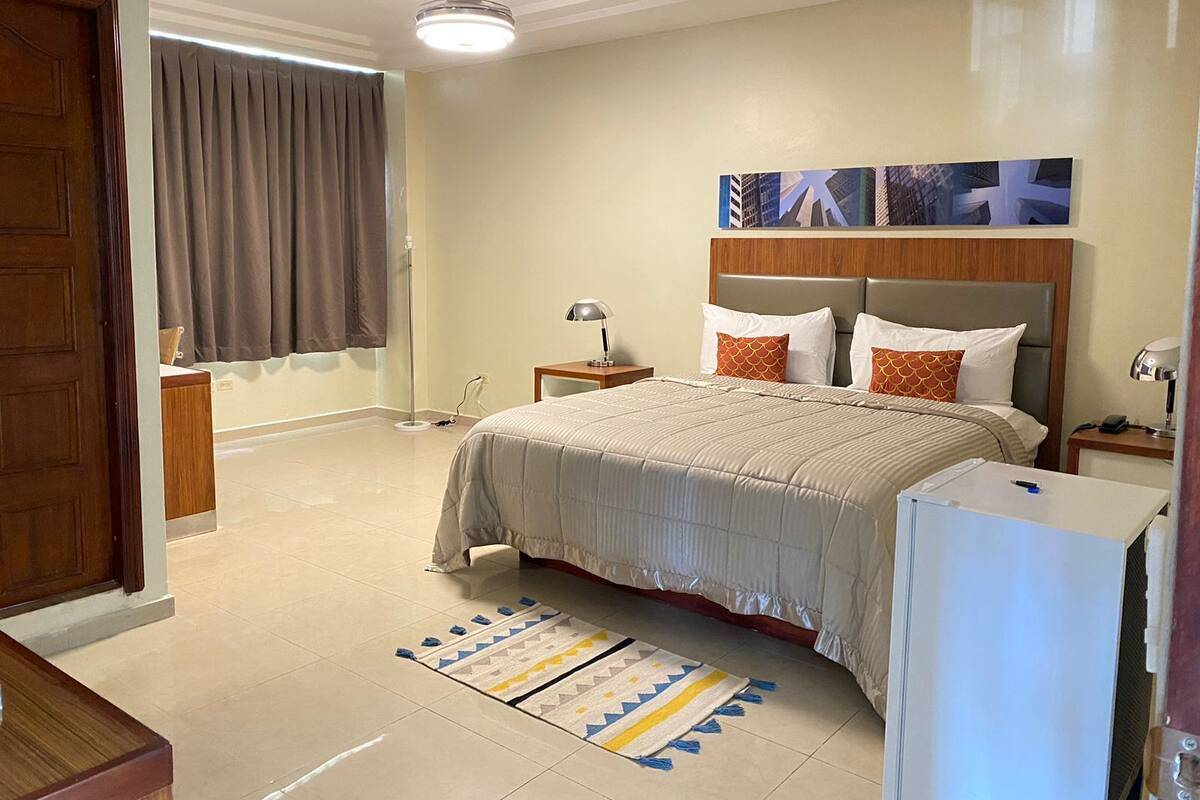 Une chambre d’hôtel comprenant un lit, une table de chevet, une lampe, un ventilateur de plafond, une fenêtre avec des rideaux et une œuvre d’art accrochée au mur.
