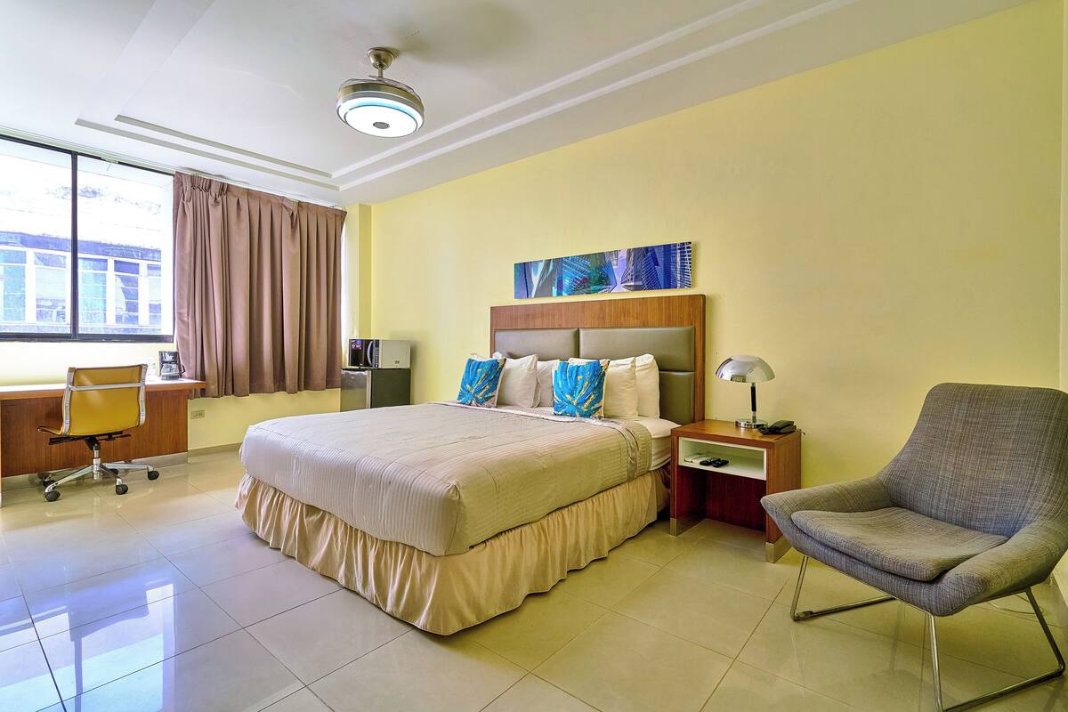 Une chambre d’hôtel avec un grand lit, un bureau avec une chaise, une petite table avec une lampe et une fenêtre avec des rideaux.