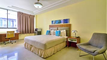 Habitación de hotel con una cama grande, un escritorio con silla, una mesita de noche con lámpara y una ventana con cortinas.