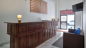 Een houten receptiebalie, twee wandlampen, een groot raam met jaloezieën, een plafondlamp en uitzicht buiten door een deur.
