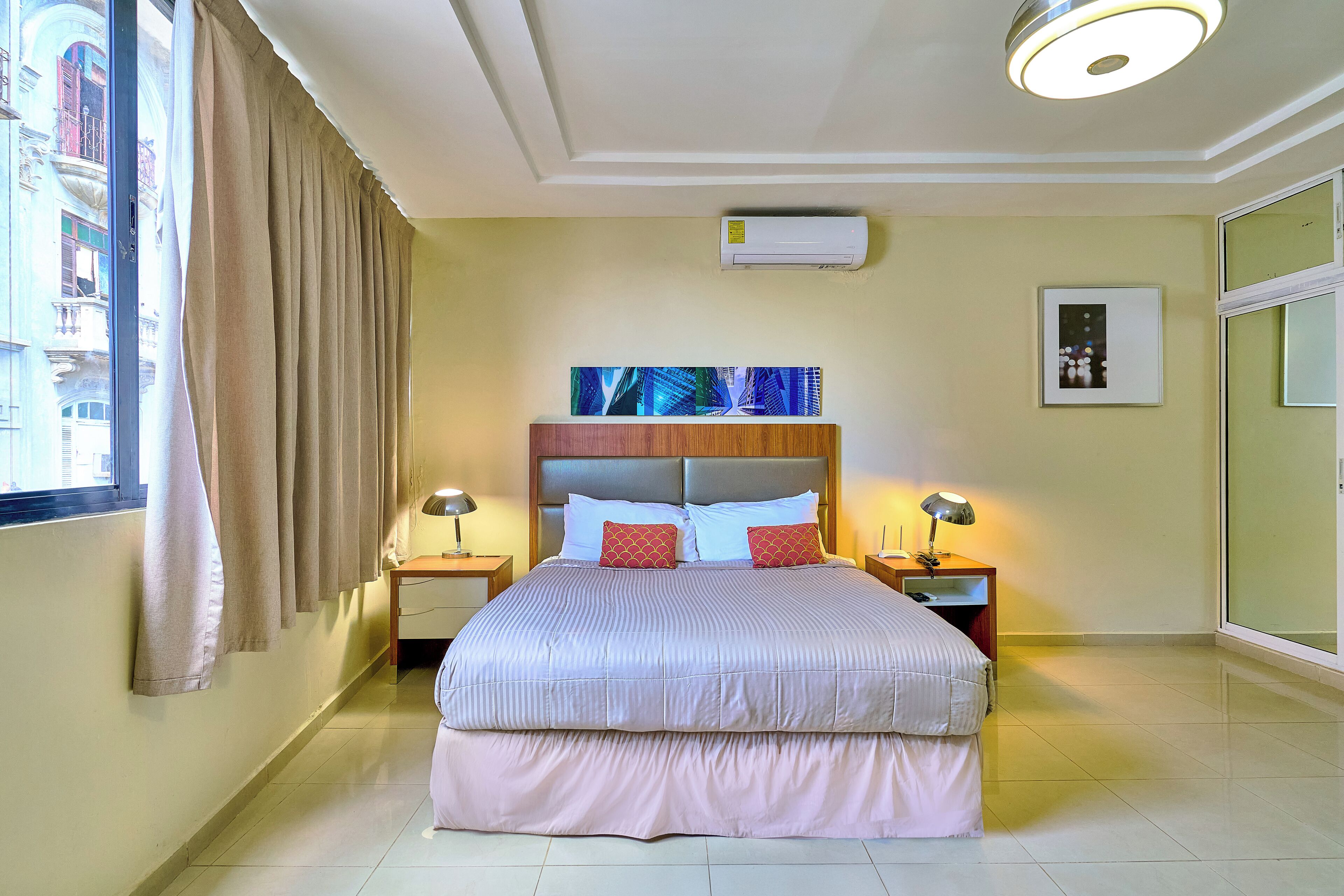 Une chambre d’hôtel avec un grand lit, deux tables de chevet avec des lampes, un luminaire au plafond, un climatiseur, un tableau encadré et une vue sur les bâtiments par la fenêtre.