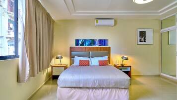 Une chambre d’hôtel avec un grand lit, deux tables de chevet avec des lampes, un luminaire au plafond, un climatiseur, un tableau encadré et une vue sur les bâtiments par la fenêtre.