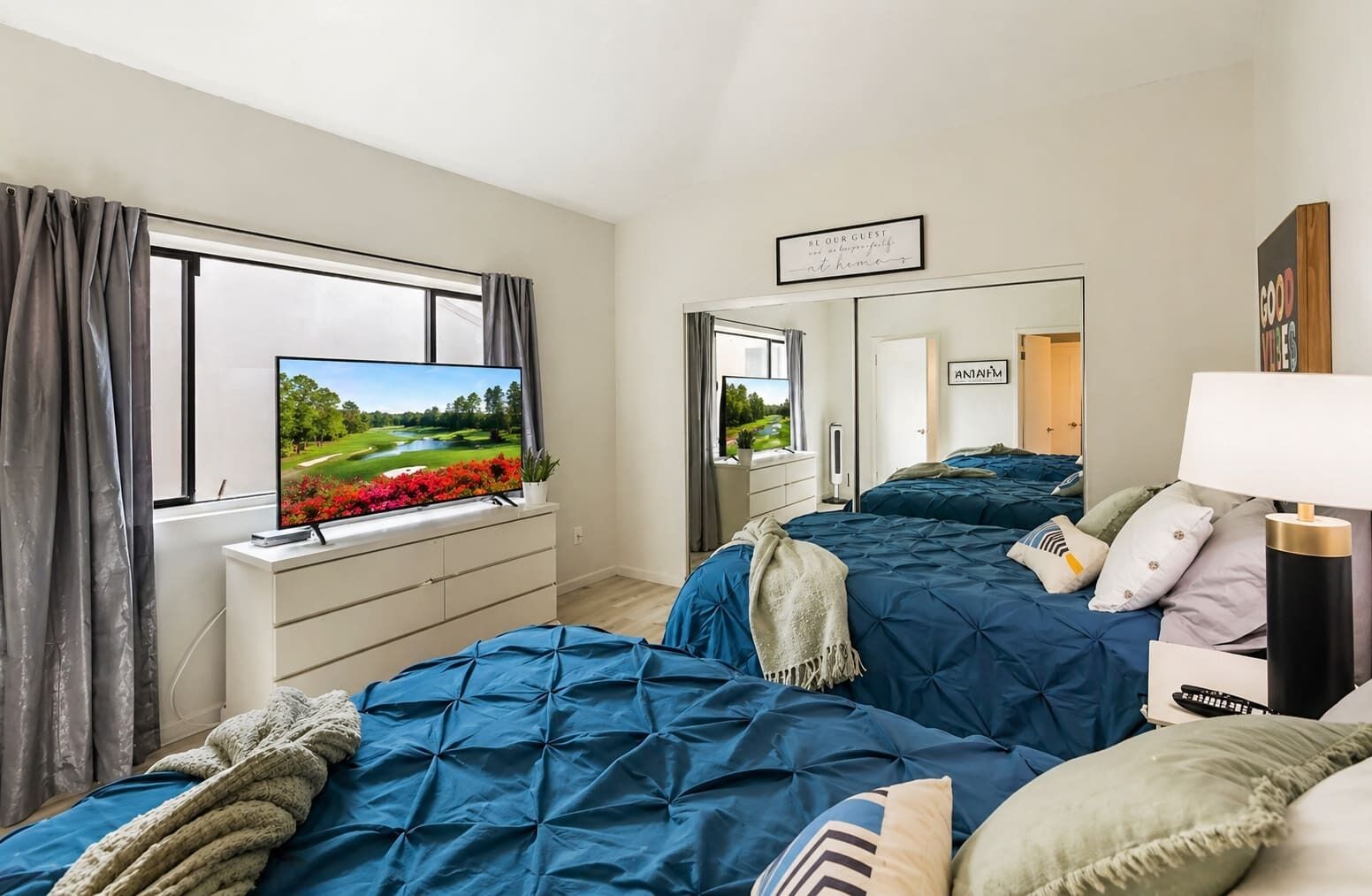 Un dormitorio con televisor sobre un tocador, cama con ropa de cama azul, un espejo y un cartel que dice 'Be Our Guest'.