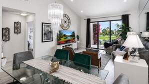 A modern living room with a glass dining table, comfortable seating, a large TV, and a view of a garden through the sliding glass door.