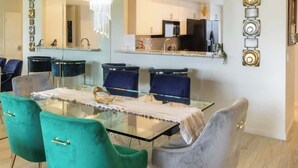 A dining area with a glass table, teal and gray chairs, a chandelier, and a kitchen in the background.