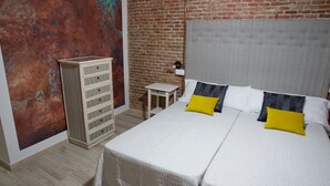 A bedroom with a brick wall, a wooden dresser, a small table, and a bed with white bedding and two pillows.
