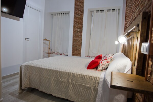 Een netjes opgemaakt bed met een beddekleed met dessin en kussens, een nachtkastje met een lamp, een houten stoel en een bakstenen muur.