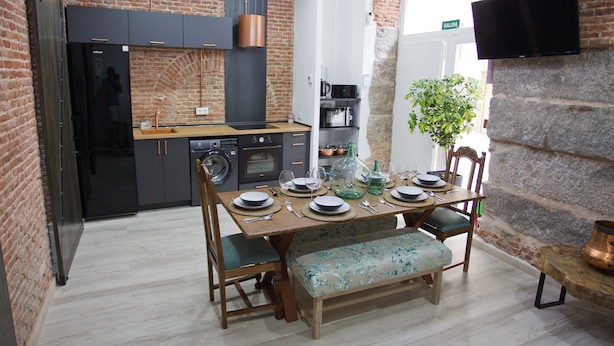 A modern kitchen with a brick wall, a dining table set for four, and a television mounted on the wall.
