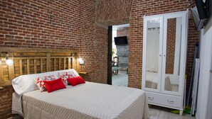 A bedroom with a brick wall, a wooden headboard, a bed with white bedding and red pillows, a wardrobe, and a mirror.