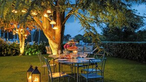 Zona pranzo all'aperto con tavoli e sedie, circondata da alberi e illuminata da luci a catena.