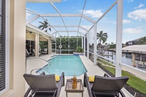 A covered outdoor swimming pool with lounge chairs and a table, surrounded by palm trees and a view of a marina. - Alluring Waterfront Home | W Exposure, Pool/Dock (Marco Island)
