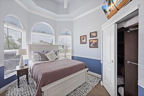 A bedroom with a bed, a nightstand, a lamp, a window with blinds, and a door. - Alluring Waterfront Home | W Exposure, Pool/Dock (Marco Island)
