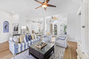 A spacious living room with a blue-striped sofa, a glass coffee table, and a ceiling fan. - Alluring Waterfront Home | W Exposure, Pool/Dock (Marco Island)