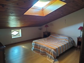 A bedroom with a sloped ceiling, a skylight, a bed with a striped bedspread, a small window, and a nightstand with a lamp.
