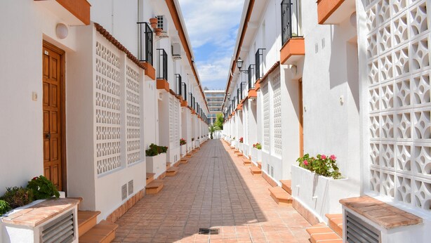 A narrow, tiled pathway flanked by white buildings with terracotta roofs, wooden doors, and decorative lattice panels. - Ola Azul Bungalow V (Benidorm)