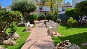 A paved pathway leading to white chairs arranged for seating, surrounded by well-maintained greenery and large rocks. - Ola Azul Bungalow V (Benidorm)