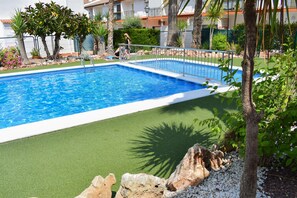 A rectangular swimming pool with a metal railing, surrounded by artificial grass, rocks, and palm trees.