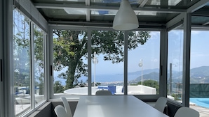 A modern dining area with a long white table and chairs, large windows offering a view of the sea and hills.