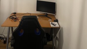 Una habitación pequeña y poco amoblada, con un escritorio, un monitor de computadora, audífonos y una silla.