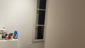 Una habitación con ventana, pared blanca, luz en el techo, una mesita con cafetera, un cuenco de fruta y un recipiente azul.