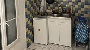Una cocina compacta con armarios blancos, un refrigerador pequeño, una estufa y un lavamanos. Hay una ventana a la izquierda y un bolso azul con lunares cerca del lavamanos.