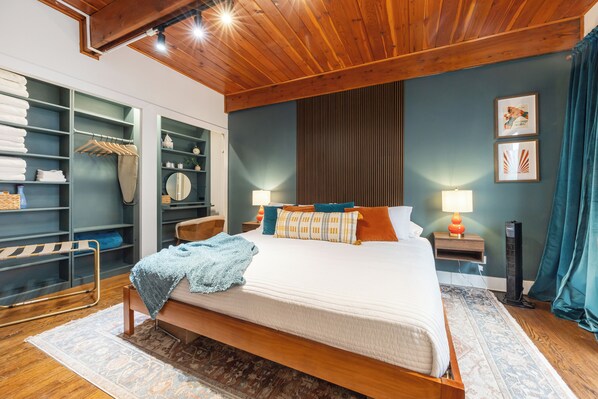 A bedroom with a wooden ceiling, a bed with a blue blanket, a nightstand with a lamp, a wardrobe, and a mirror. - Lincoln Park 3BR Townhome • Rooftop • King Suite • Sleeps 10 (Chicago)