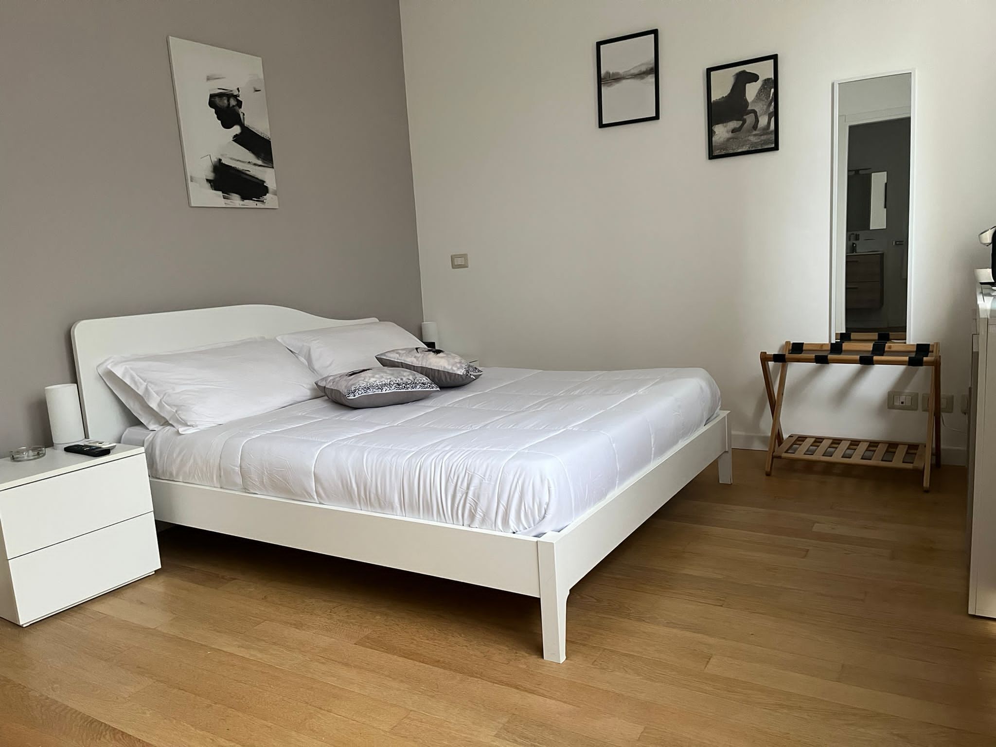 Un dormitorio ordenado con una cama blanca, una mesita de noche, una mesita auxiliar de madera y un espejo.