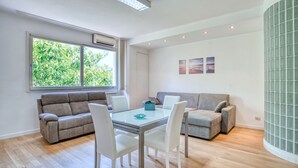 Ein modernes Wohnzimmer mit einem Glastisch, weißen Stühlen, einem grauen Sofa und einem grauen Sessel. Ein Fenster bietet Blick auf Grünfläche.