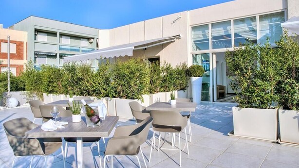 Ein möblierter Patio-Bereich mit Tischen und Stühlen, umgeben von viel Grün und einem modernen Gebäude im Hintergrund.