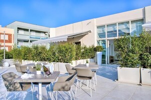 Un patio al aire libre con mesas y sillas, rodeado de follaje y un edificio moderno al fondo.