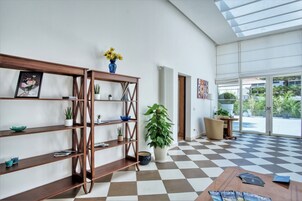 Amplia habitación con suelo de cuadros, estanterías de madera con plantas, una planta grande en maceta y vistas a través de un ventanal.