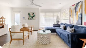 Ein Wohnzimmer mit einem blauen Sofa, einem runden Couchtisch und einem weißen Sessel. Eine Tür und ein Fenster mit Jalousien sind ebenfalls vorhanden.