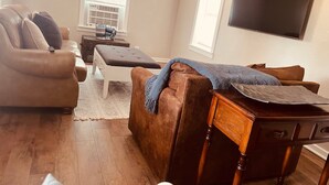 A living room with a brown leather sofa, a white ottoman, a wooden side table, and a flat-screen TV mounted on the wall.