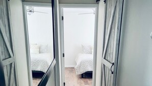 A narrow hallway with a wooden floor leading to two bedrooms with white bedding and a ceiling fan.