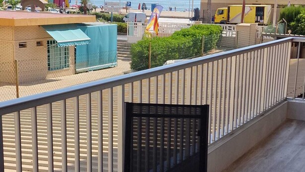 Un balcón con una mesa y dos platos, con vista a una playa con palmeras y un estacionamiento.