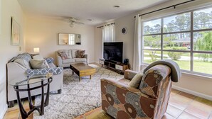 A living room with a sofa, armchair, coffee table, TV, and a large window with a view of greenery. - Pools Paradise: 4  BR, 2  BA House in Wilmington, Sleeps 8 (Wilmington)