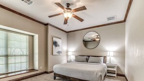 Une chambre avec un grand lit, un ventilateur de plafond, une fenêtre avec des stores, une table de chevet avec une lampe et un miroir fixé au mur.
