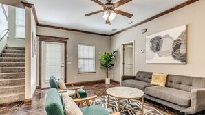 Un salon spacieux avec un canapé gris, des fauteuils verts et une table basse. On peut voir un ventilateur de plafond et un grand tableau abstrait.