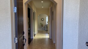 Un pasillo con puerta de madera, paredes color beige y suelo de baldosas que conduce a una habitación con ventana circular y una planta.