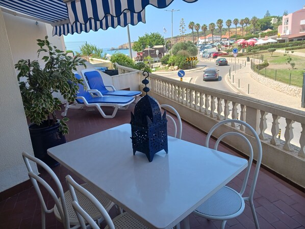 A balcony with a table and chairs, a potted plant, and a view of the sea and buildings.