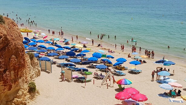 Een strand met talloze parasols en ligbedden, een groot cruiseschip op de achtergrond en een rotsachtige klif aan de linkerkant.