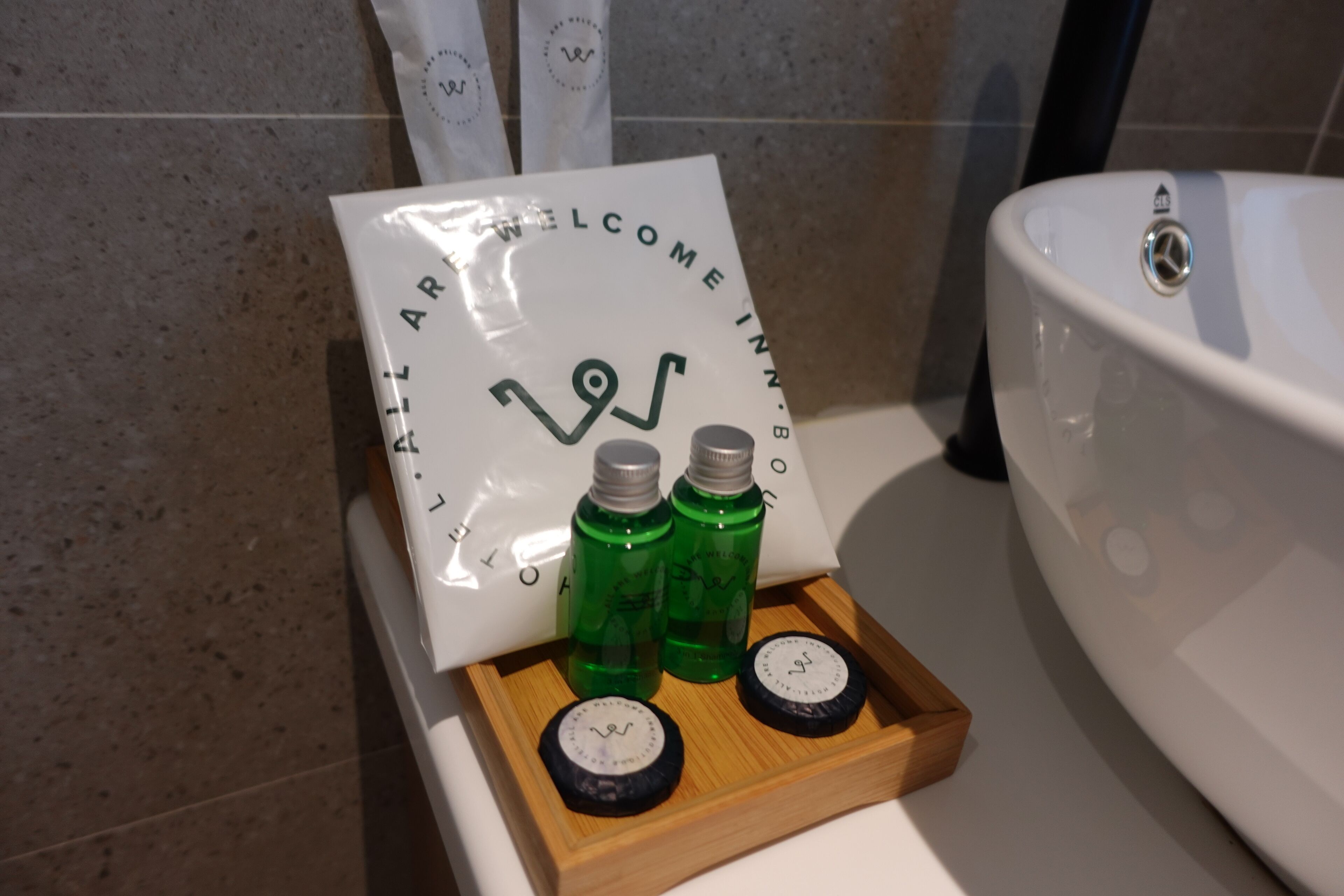 A bathroom with a white sink, a wooden tray holding two green bottles and two black circular objects, and a sign with a logo and text.