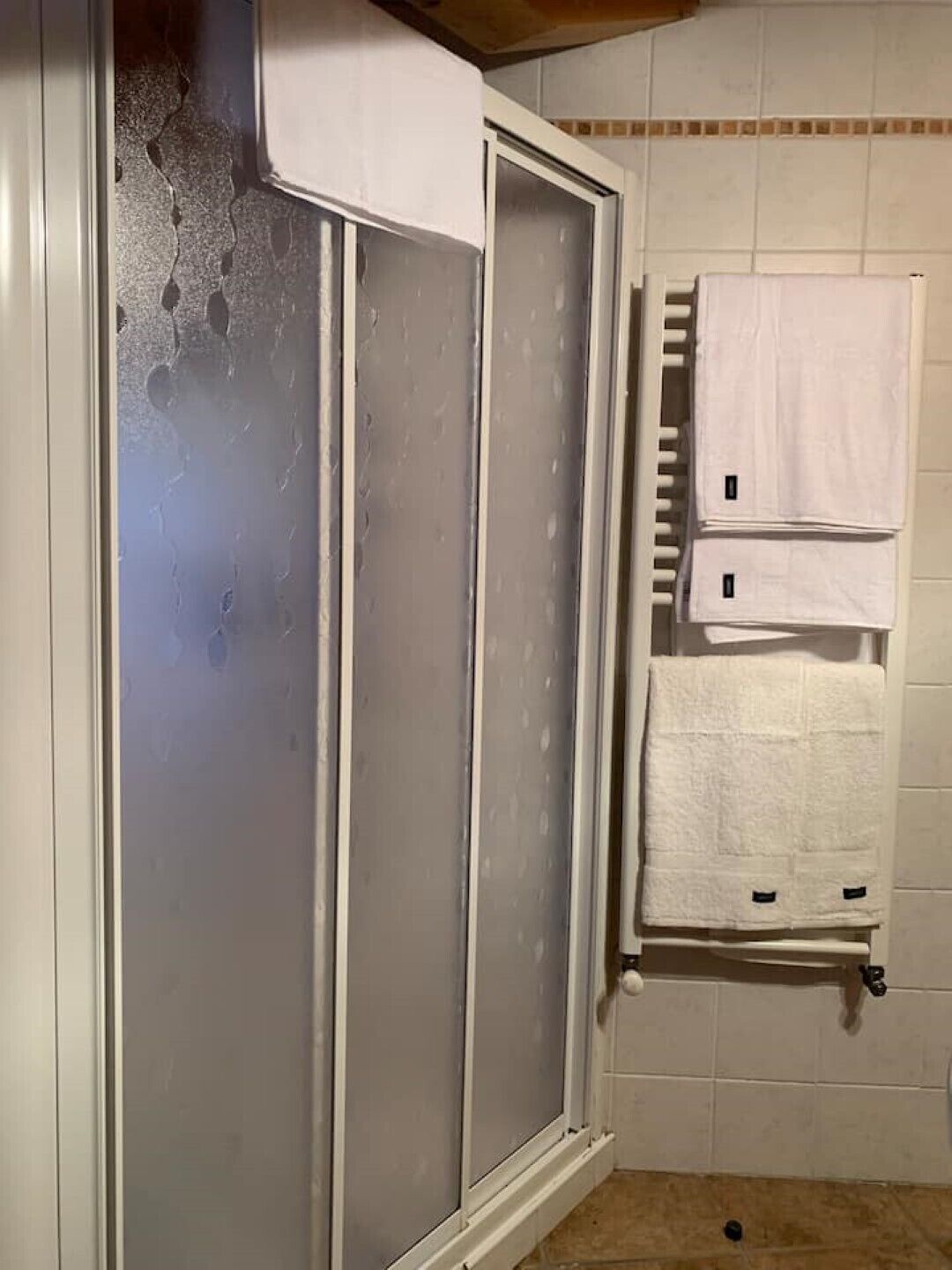 A bathroom with a frosted glass shower enclosure, a towel rack with two towels, and a tiled wall.