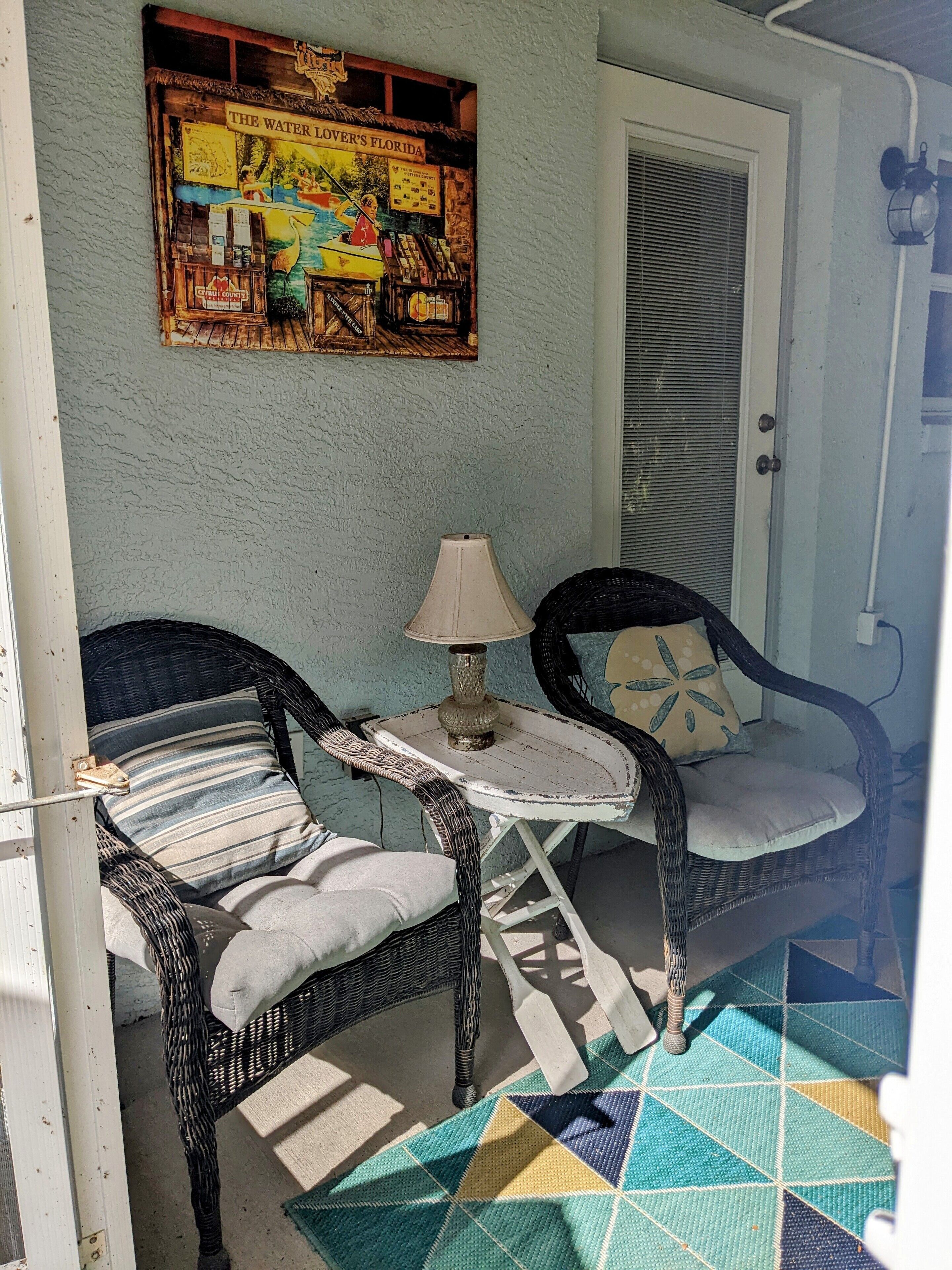 A room with two wicker chairs, a small table with a lamp, a colorful rug, and a wall-mounted poster.