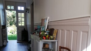 Un passadís amb rajoles de terra estampades, una cadira de vimet, una petita estant amb objectes decoratius i una porta que dona a l'exterior.