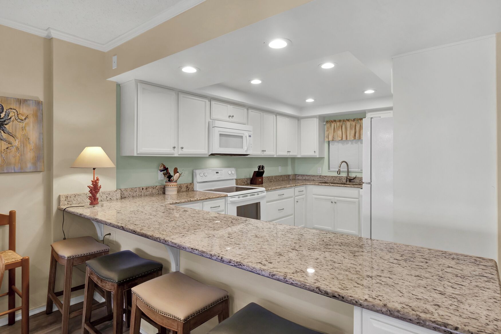 A kitchen with white cabinets, a granite countertop, a stove, and a sink. There are three stools and a table with a lamp.