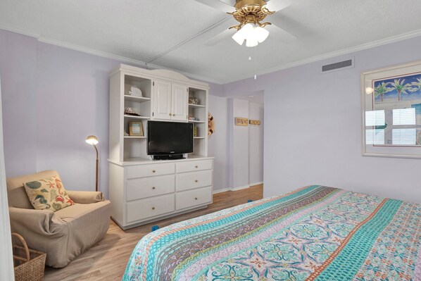 A bedroom with a bed, a TV on a white cabinet, a chair, and a ceiling fan.